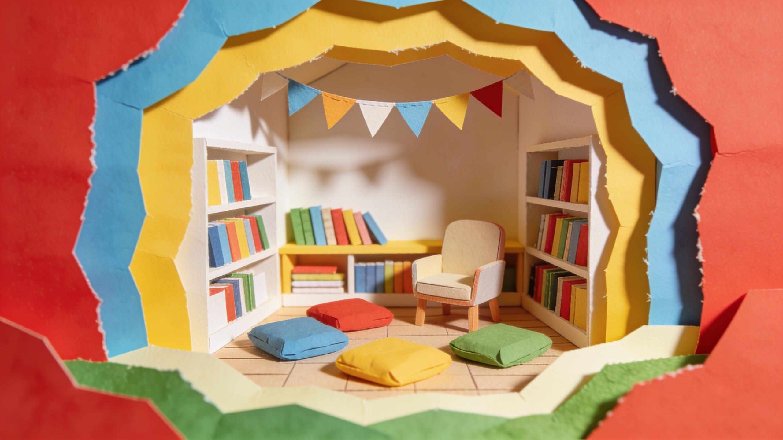 A paper-craft reading corner with cushions on the floor, a small armchair and shelves of colourful books under bunting